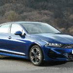 A blue 2021 Kia Optima is parked in a parking lot with mountains in the distance.