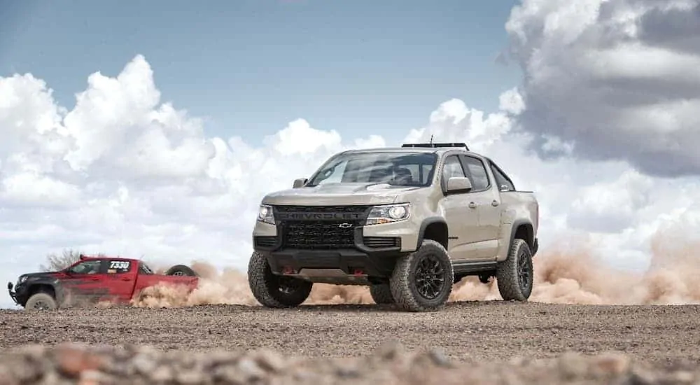 A tan 2021 Chevy Colorado ZR2 is parked in the desert while a red one races past.