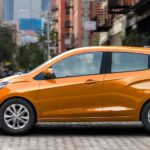 An orange 2020 Chevy Spark is driving on a city street after leaving a Chevy dealership.