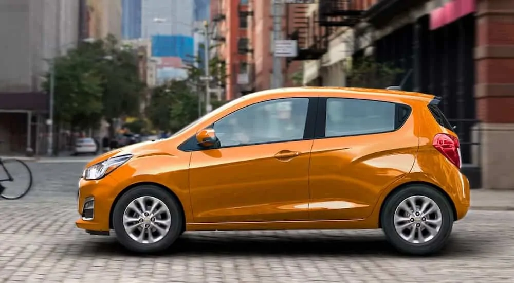 An orange 2020 Chevy Spark is driving on a city street after leaving a Chevy dealership.