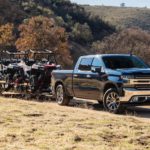 A dark grey 2020 Chevy Silverado is towing side by sides on a dirt road.