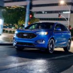 A blue 2020 Ford Edge is driving on a city street at night.