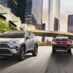 A red 2020 Toyota RAV4 is driving behind a silver one on a multi-lane city street.