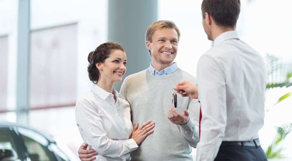 A salesman is handing keys to a happy couple inside a Chevy dealership showroom.