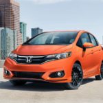 An orange 2020 Honda Fit is parked on a rooftop parking garage with city buildings behind it.