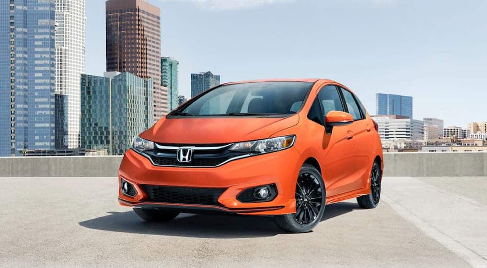 An orange 2020 Honda Fit is parked on a rooftop parking garage with city buildings behind it.