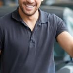 A man is holding keys while leaning on a black car after seeing a dealership ad to 'Sell Us Your Car.'