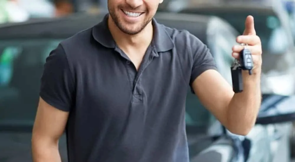 A man is holding keys while leaning on a black car after seeing a dealership ad to 'Sell Us Your Car.'