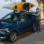 A couple is loading kayaks on the roof of a blue 2020 Buick Encore GX next to a lake.