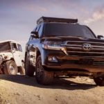 A black 2020 Toyota Land Cruiser is parked on a dirt road in front of a vintage white model.