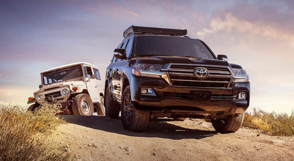 A black 2020 Toyota Land Cruiser is parked on a dirt road in front of a vintage white model.