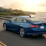 A blue 2020 Chevy Malibu is driving along an ocean bay.