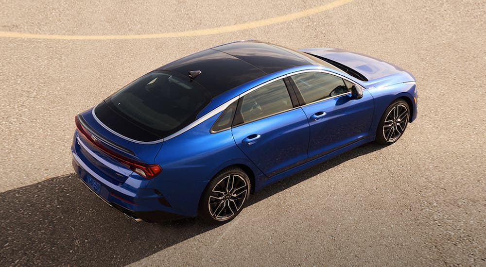 A blue 2021 Kia K5 is shown from above while parked in an empty lot.