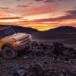 A yellow 2021 Ford Bronco 2 door is parked on rocks in front of mountains and a vibrant orange sunset.