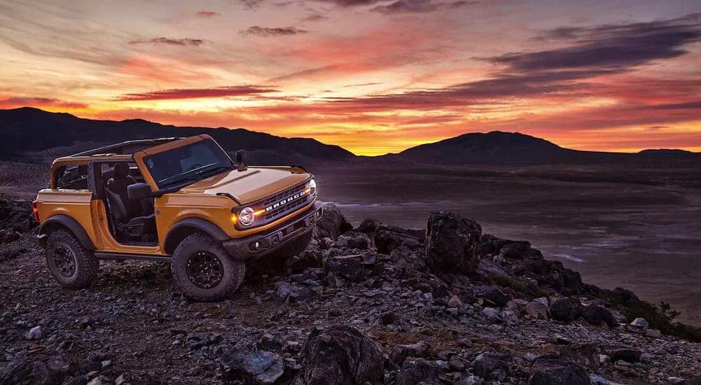 A yellow 2021 Ford Bronco 2 door is parked on rocks in front of mountains and a vibrant orange sunset.