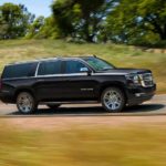 A popular Chevy SUV, a black 2020 Chevy Suburban, is driving on a rural highway and shown from the side.