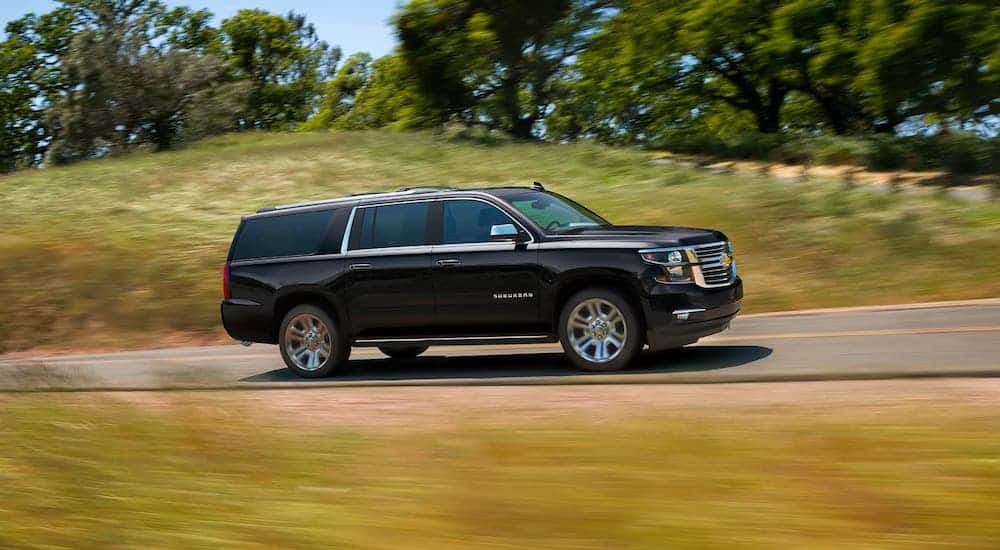 A popular Chevy SUV, a black 2020 Chevy Suburban, is driving on a rural highway and shown from the side.