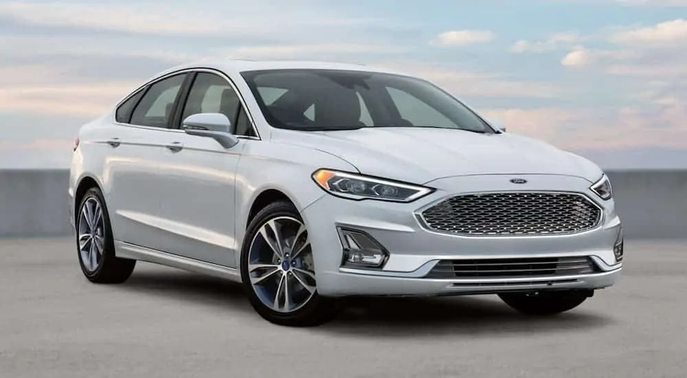 A white 2020 Ford Fusion is parked in front of a low cement wall and blue, cloudy sky after winning the 2020 Ford Fusion vs 2020 Mazda 6 comparison.