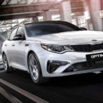 A white 2020 Kia Optima is driving under a bridge.