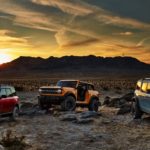 In a comparison of the 2021 Ford Bronco vs 2021 Ford Bronco Sport, three models are parked in front of a sunset behind mountains.