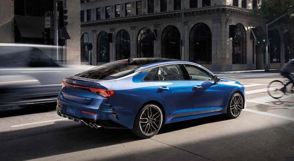 A blue 2021 Kia K5 is stopped at an intersection on a busy city street.