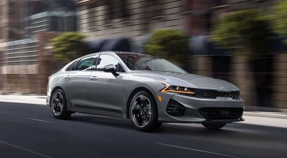 A grey 2021 Kia K5 is driving down a city street after winning the 2021 Kia K5 vs 2020 Kia Optima comparison.