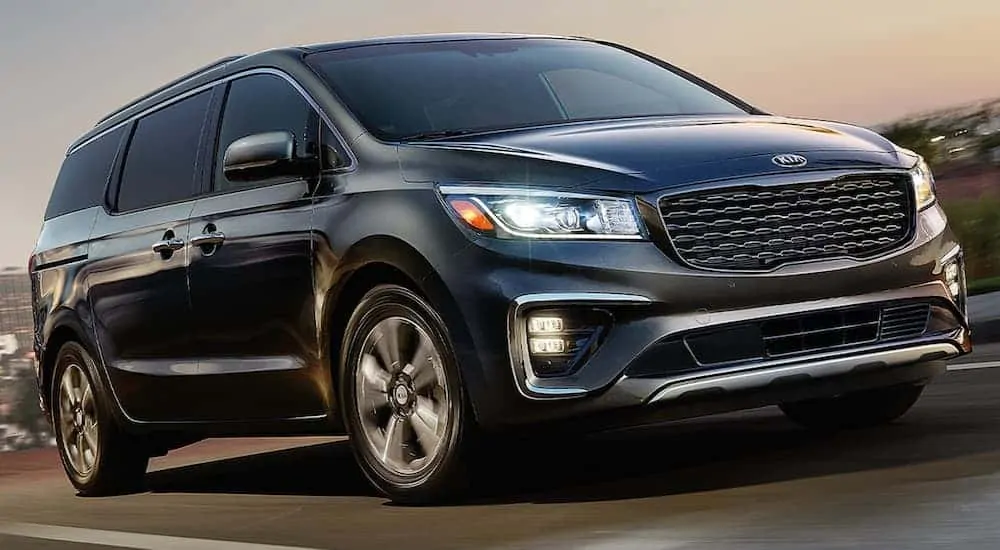 A grey 2021 Kia Sedona is driving past a blurred city at dusk.