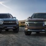 A grey 2021 Chevy Tahoe is parked next to a brown Suburban on a rural road.