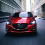 A red 2014 Mazda3 from a used car dealership near you is driving on a city street and shown from the front.