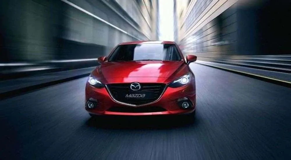 A red 2014 Mazda3 from a used car dealership near you is driving on a city street and shown from the front.