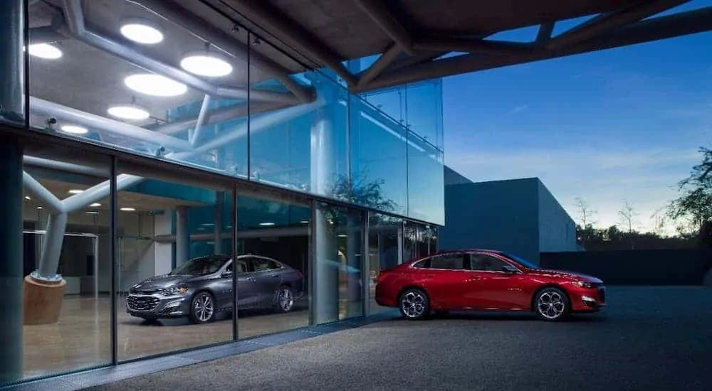 A red 2020 Chevrolet Malibu parked in front of a glass building with its mirrored image in silver on the building.