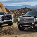 A white and a black 2020 GMC Sierra 2500 are parked on dirt in front of mountains.