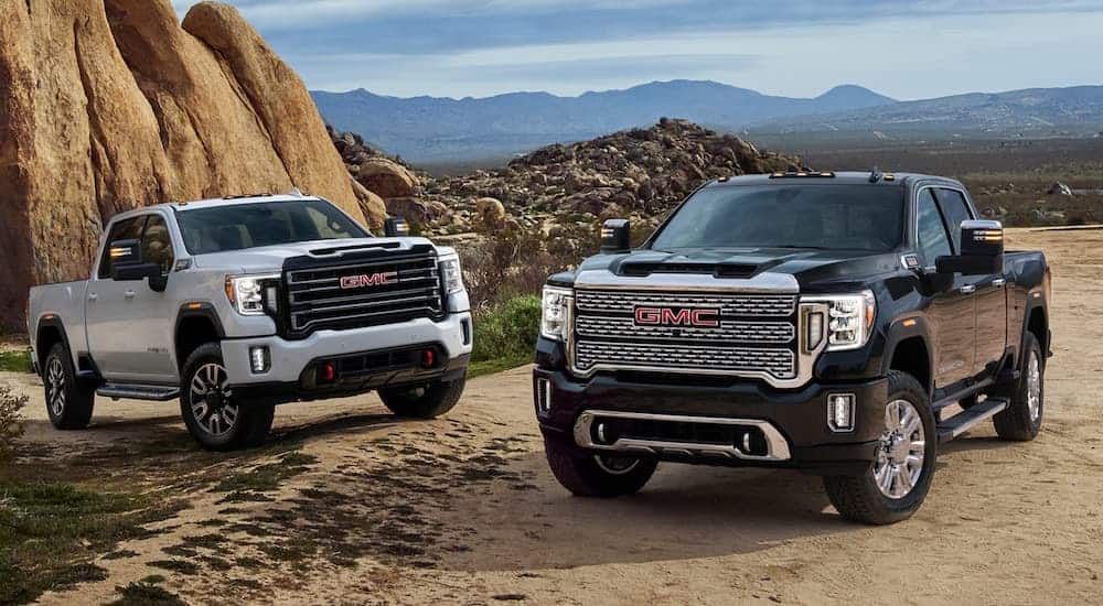 A white and a black 2020 GMC Sierra 2500 are parked on dirt in front of mountains.