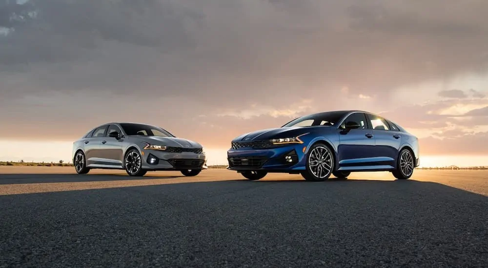 Two 2021 KIA K5's one blue, one grey, are parked on the pavement with the sun setting in the background.