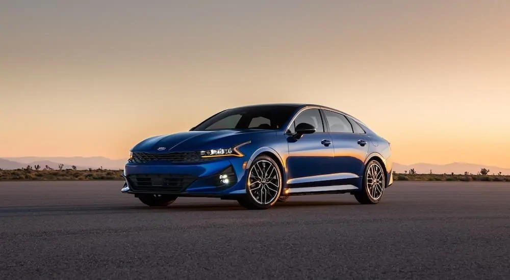 A blue 2021 Kia K5 from a Kia dealership near me is parked on the asphalt at dusk.