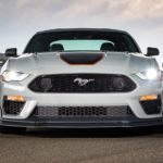 A white 2021 Ford Mustang Mach 1 is parked at the track with its headlights illuminated.