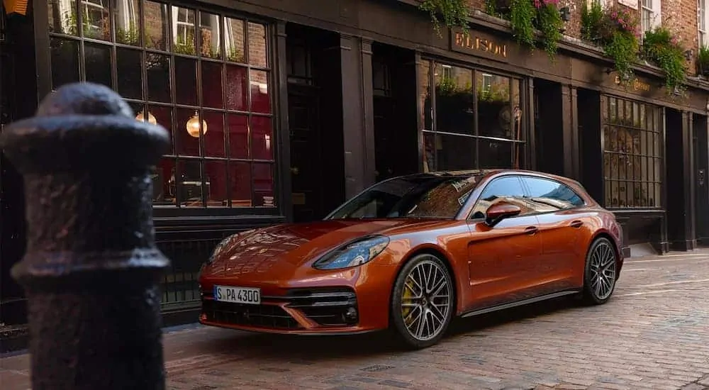 A orange 2021 Porsche Panamera is parked on a brick road near a store front.