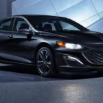 A black 2021 Chevy Malibu is parked in front of a modern building with the headlights on.