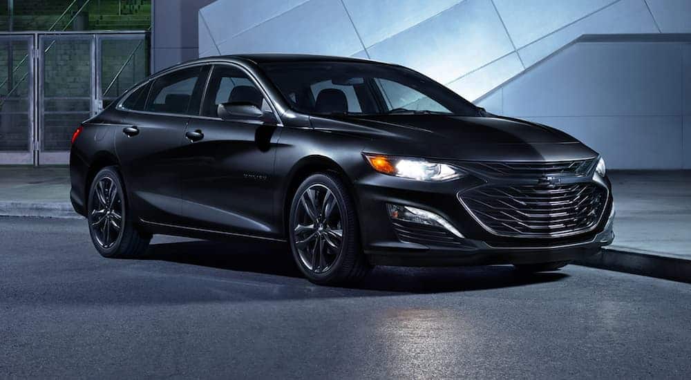 A black 2021 Chevy Malibu is parked in front of a modern building with the headlights on.