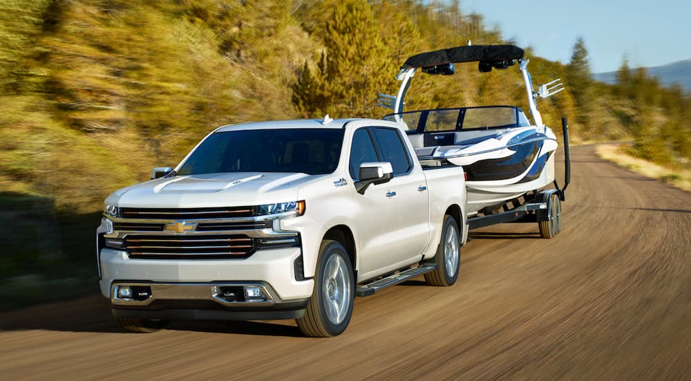 A white 2021 Chevy Silverado 1500 is towing a boat on a winding road past a lake.