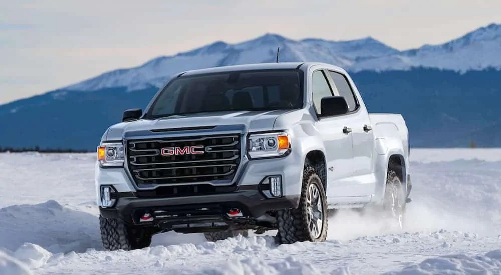 A white 2021 GMC Canyon AT4 is driving through the snow with mountains in the background.
