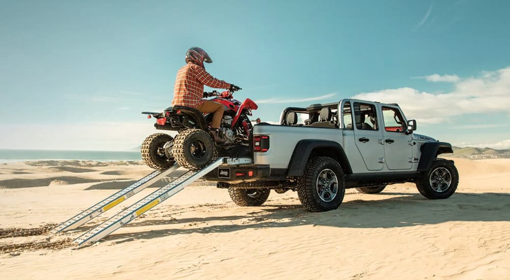 A person is riding a four wheeler into the bed of a silver 2021 Jeep Gladiator on sand at the ocean.