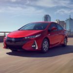 A red 2021 Toyota Prius Prime is driving on a highway away from a city.