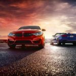 A red 2021 BMW M4 Coupe and a blue 2021 BMW M4 Convertible are parked on wet pavement at sunrise.