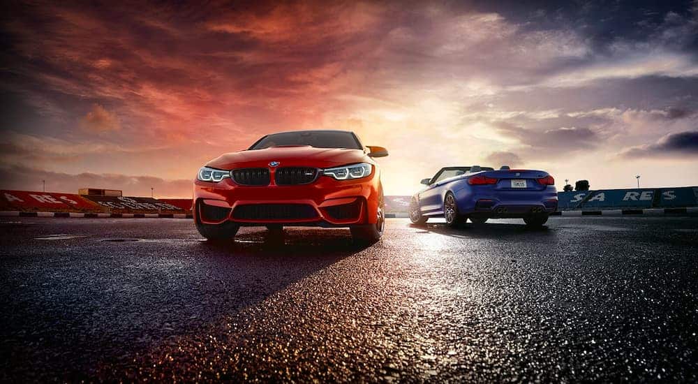 A red 2021 BMW M4 Coupe and a blue 2021 BMW M4 Convertible are parked on wet pavement at sunrise.