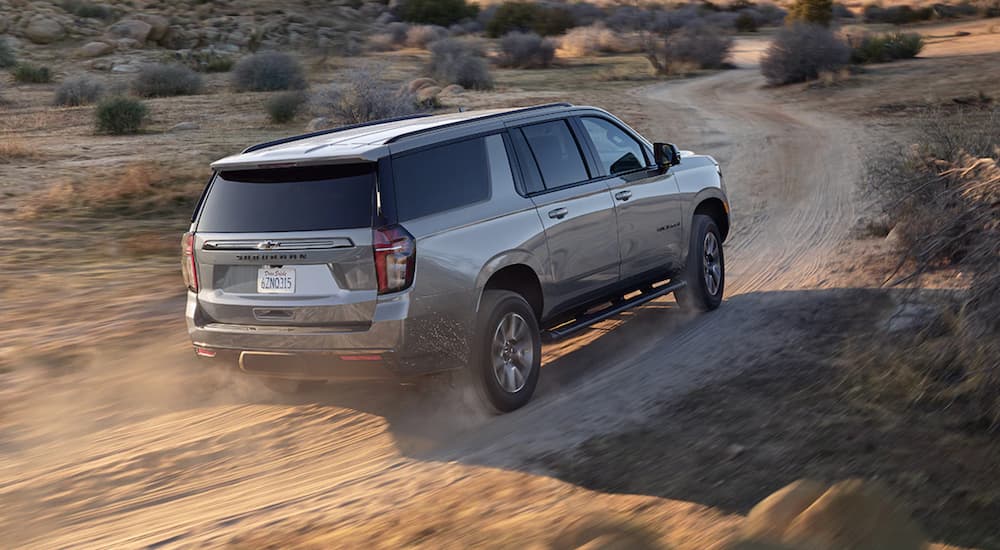 A grey 2021 Chevy Suburban is driving on a dirt road past shrubs.