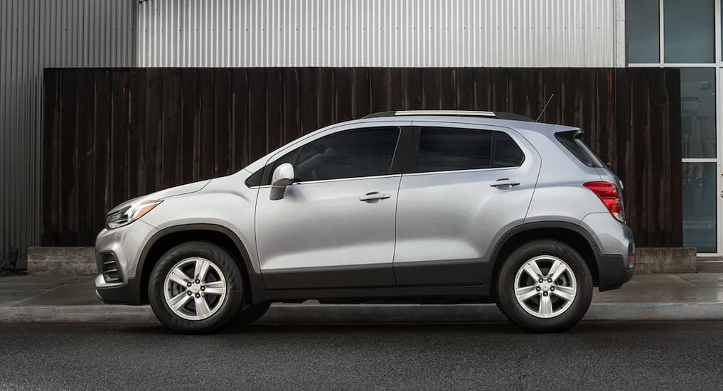 The smallest Chevy SUV, a silver 2021 Chevy Trax, is shown from the side in front of a grey metal building.