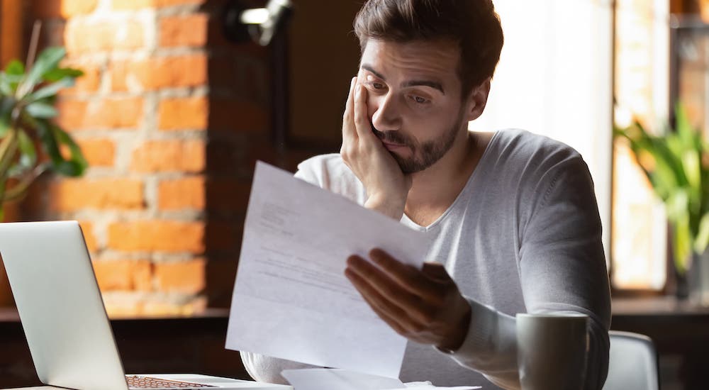 A man is in anguish while reading a piece of paper in front of a laptop.