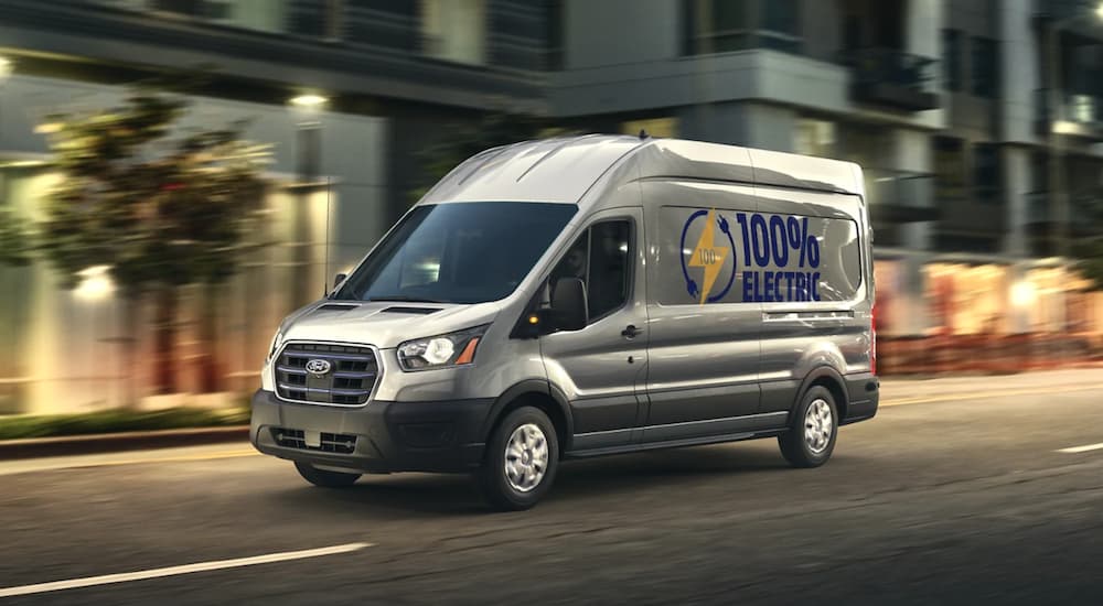 A silver 2022 Ford E-Transit is driving on a city street at dusk after leaving a Ford commercial vehicle dealer.