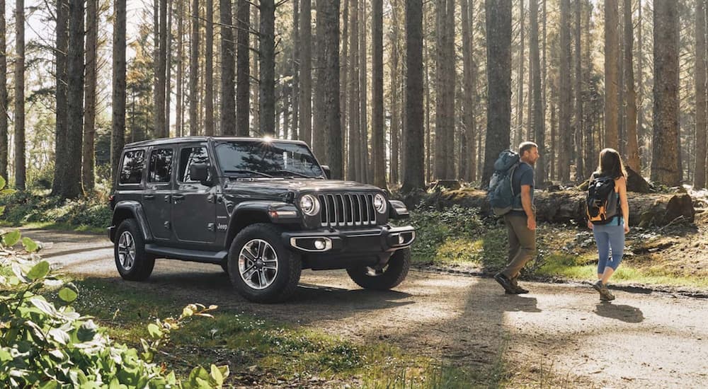 A couple is walking away from a grey 2020 Jeep Wrangler Unlimited in the woods with back packs.
